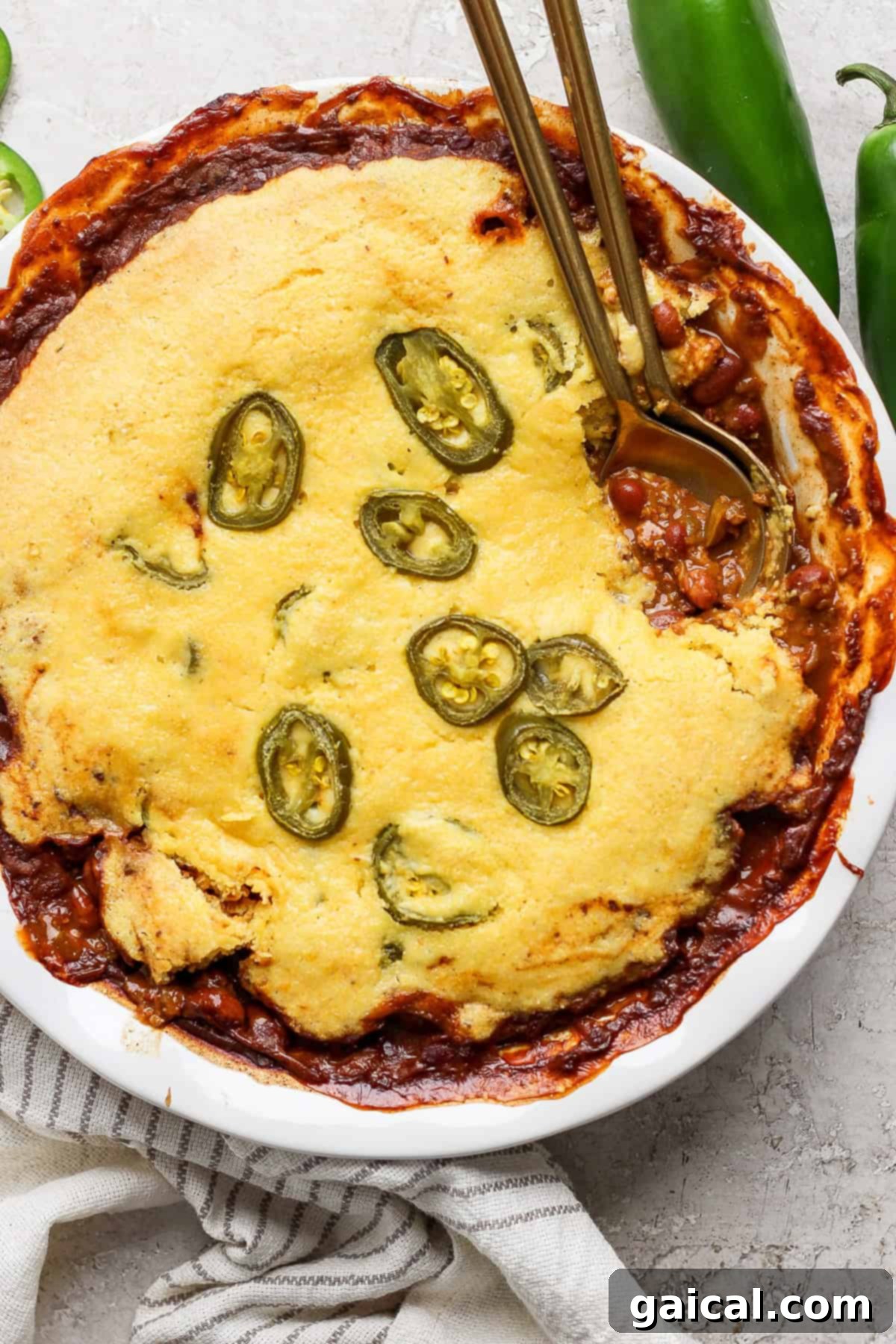 Effortless Chili Pot Bake 2 A dish of cornbread chili pie topped with sliced jalapeños, served in a white pie dish, with two gold-colored spoons placed on top, set on a light-colored surface, inviting diners to dig in.