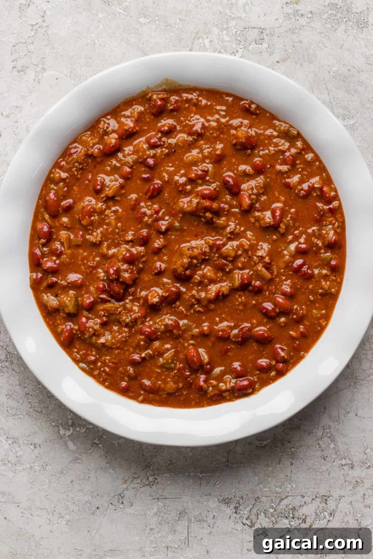 Effortless Chili Pot Bake 3 A white bowl filled with chili, containing ground meat, beans, and a thick tomato-based sauce, placed on a textured gray surface, showcasing the rich, hearty base for the pot pie.