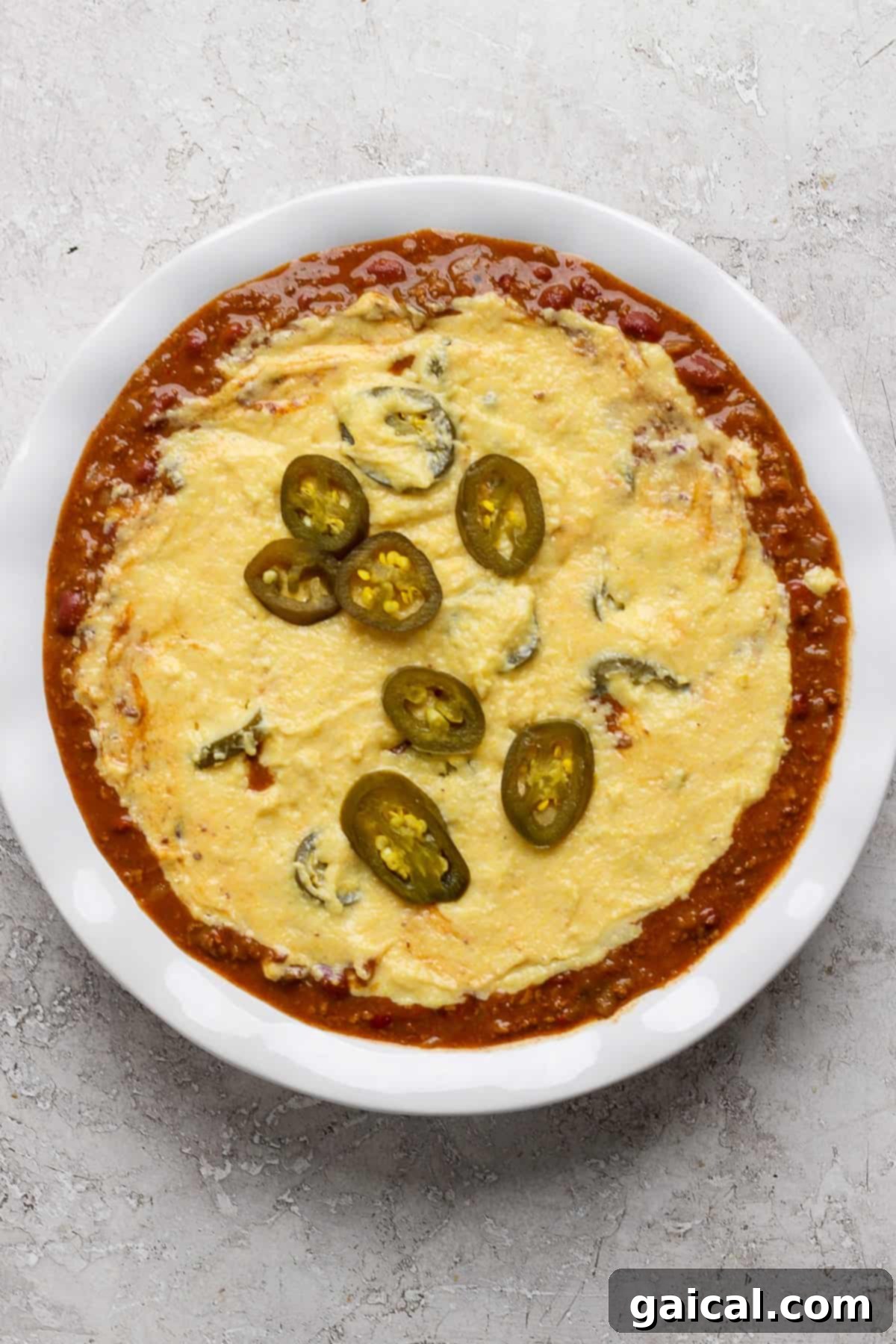 Effortless Chili Pot Bake 4 A white dish filled with melted cheese topped with sliced jalapeños, surrounded by a red tomato-based sauce, suggesting delicious add-ins for the chili pot pie.