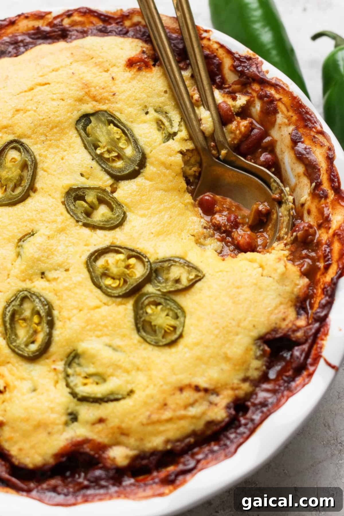 Effortless Chili Pot Bake 5 A baked dish in a white pie dish features a golden cornbread topping with sliced jalapeños, partially revealing a chili filling underneath. Two metal spoons are placed in the dish, ready for serving.