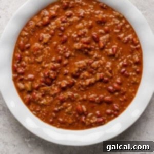 Effortless Chili Pot Bake 7 A white bowl filled with chili, containing ground meat, beans, and a thick tomato-based sauce, placed on a textured gray surface.