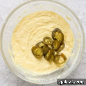 Effortless Chili Pot Bake 8 A bowl containing a creamy mixture with several slices of jalapeños placed on top. The bowl is set on a textured light-colored surface.