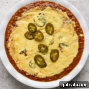 Effortless Chili Pot Bake 9 A white dish filled with melted cheese topped with sliced jalapeños, surrounded by a red tomato-based sauce.