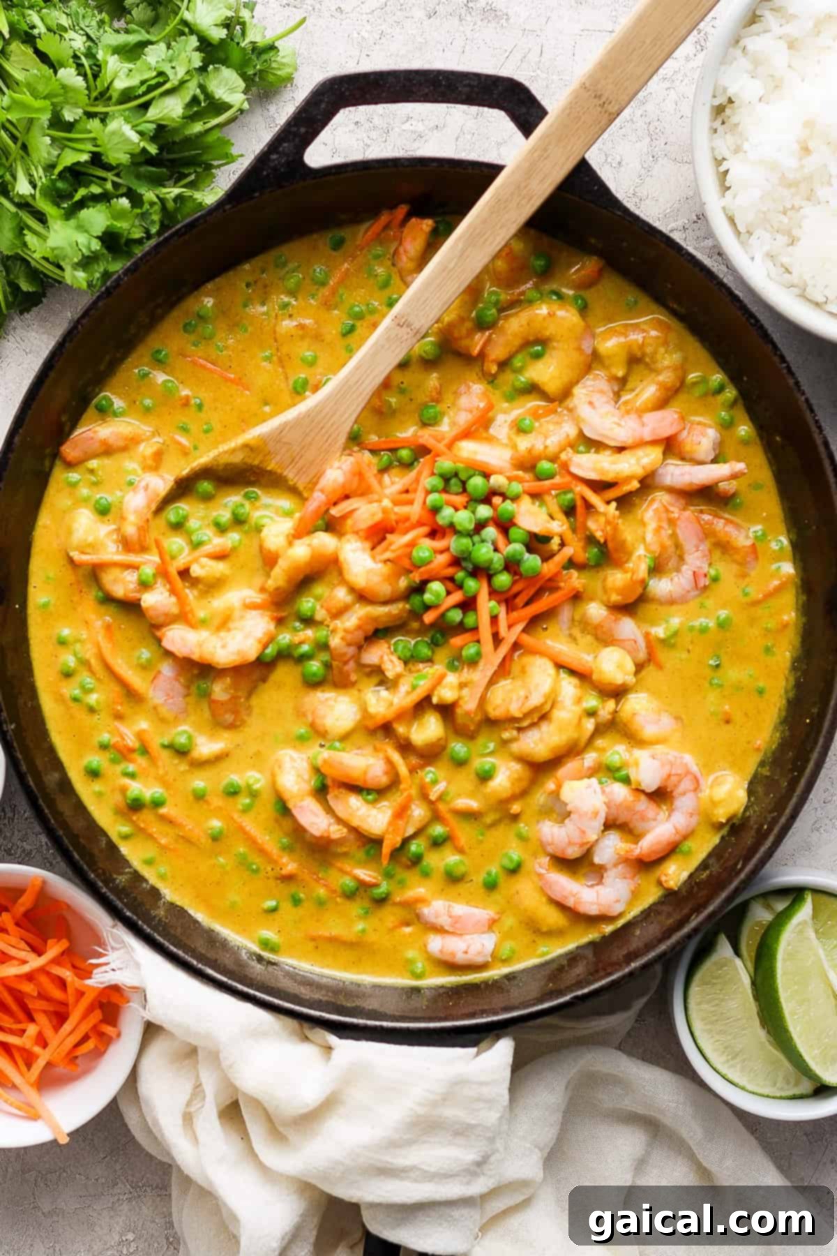 A skillet filled with shrimp and vegetable curry, garnished with green peas and served with sides of rice, lime, and fresh herbs.