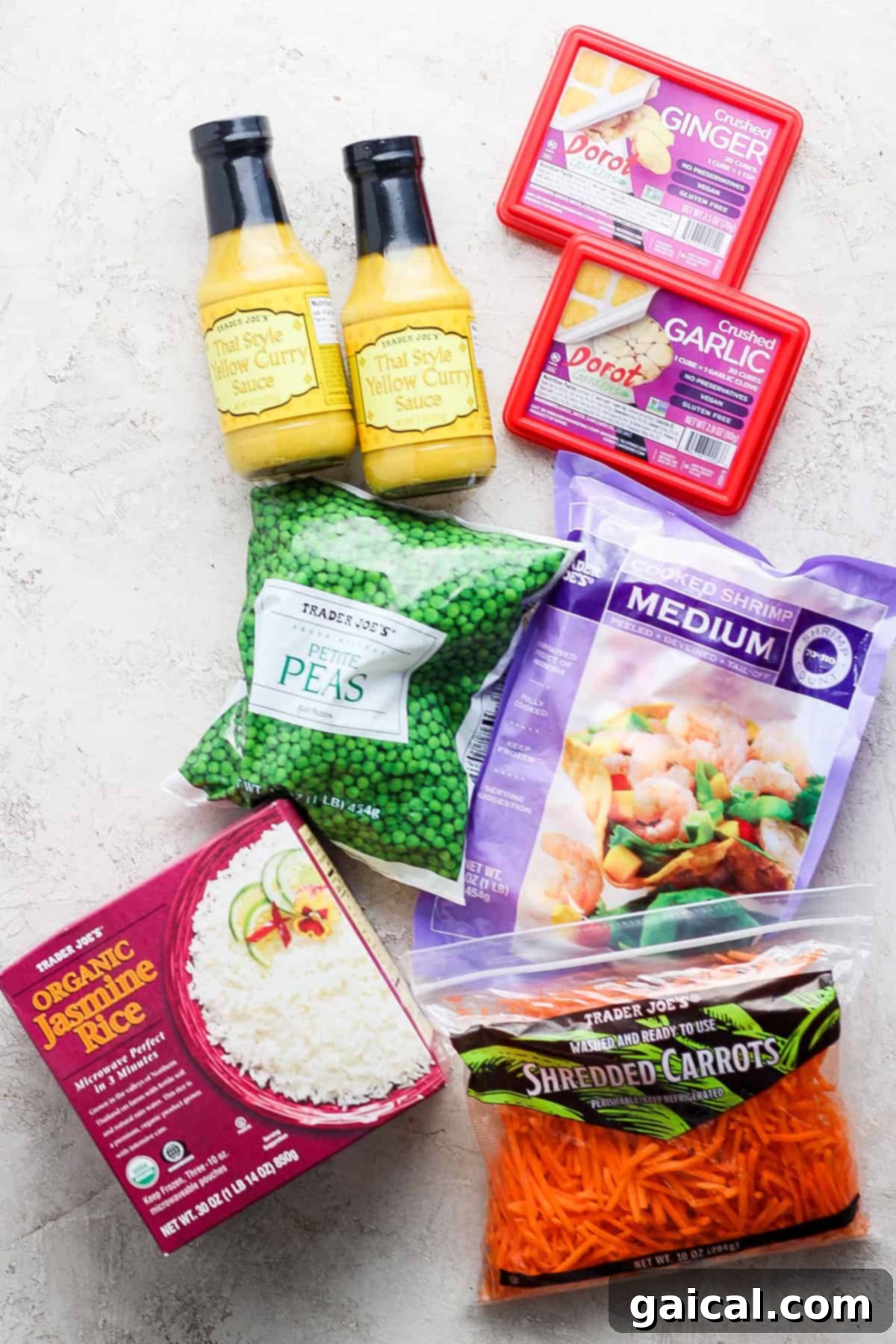 A collection of various food items, including bottles of thai yellow curry sauce, a box of ginger, packaged garlic, organic jasmine rice, frozen peas, and shredded carrots, arranged on a light textured surface.