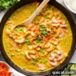 A wooden spoon is stirred into a skillet containing a shrimp curry with green peas and shredded carrots, accompanied by a side of white rice and lime wedges.