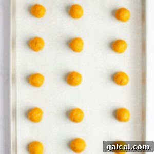 A baking tray with 15 evenly spaced, round, orange dough balls placed on a parchment-lined surface.