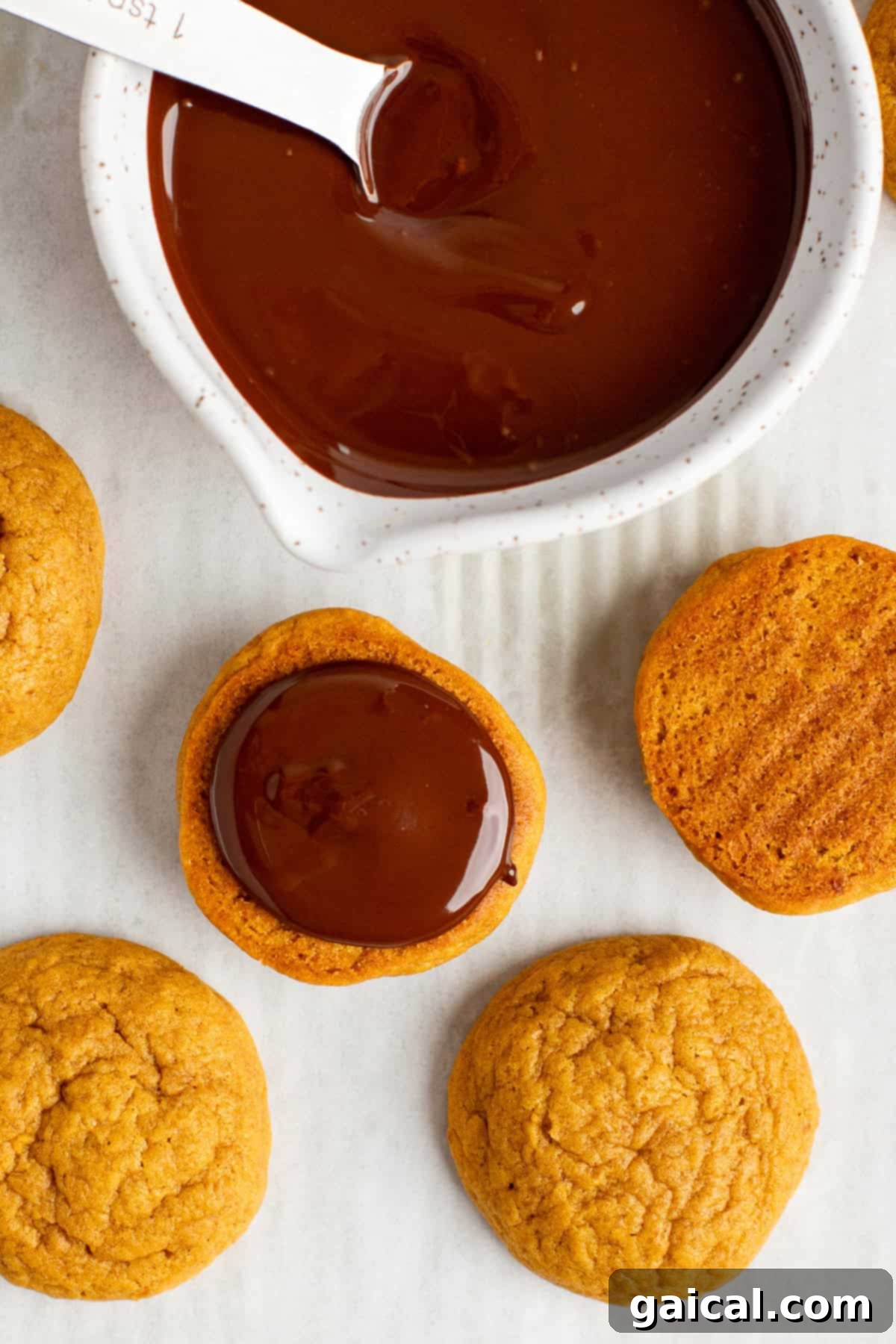 A chocolate dipped pumpkin cookie being carefully dipped into a bowl of melted chocolate, with more un-dipped cookies waiting on a white surface.