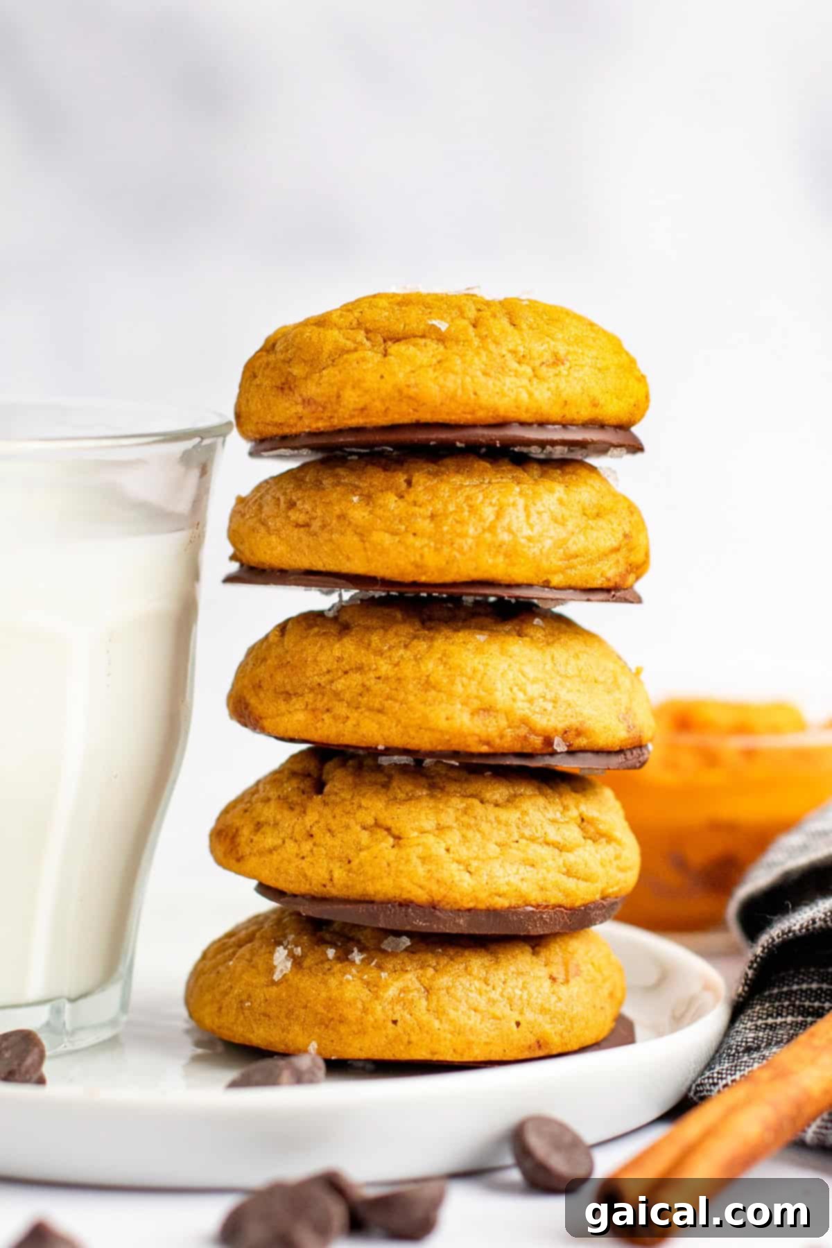 A stack of five pumpkin cookies, some with chocolate dip, elegantly arranged on a white plate. A glass of milk and a folded cloth napkin sit nearby, with chocolate chips scattered around, creating a cozy and inviting scene.