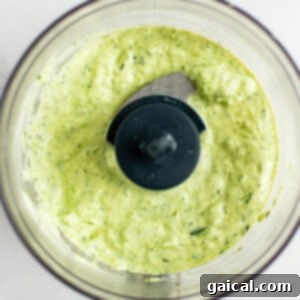 A bowl of green and white sauce sits inside a food processor with a blade in the center, illustrating the dressing preparation.