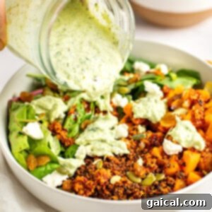 A hand pouring green herb dressing from a jar onto a bowl containing quinoa, roasted vegetables, greens, seeds, and crumbled cheese with another bowl of roasted vegetables in the background.