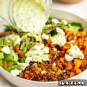 A close-up of a salad in a white bowl with quinoa, mixed greens, pumpkin seeds, raisins, and crumbled cheese, being topped with a creamy green dressing poured from a jar.