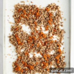 A baking tray covered with parchment paper holds a single layer of evenly spread, mixed quinoa with a toasted appearance.