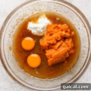A clear glass bowl containing a mixture of ingredients including two raw eggs, pumpkin puree, white yogurt, and a brown liquid, is placed on a textured white surface.
