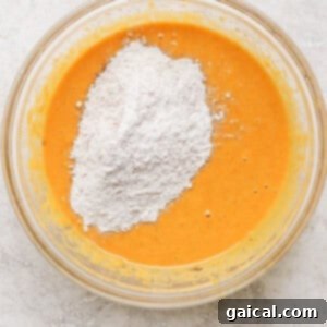 A clear glass bowl containing an orange batter with a heap of white flour added on top, placed on a marble surface.