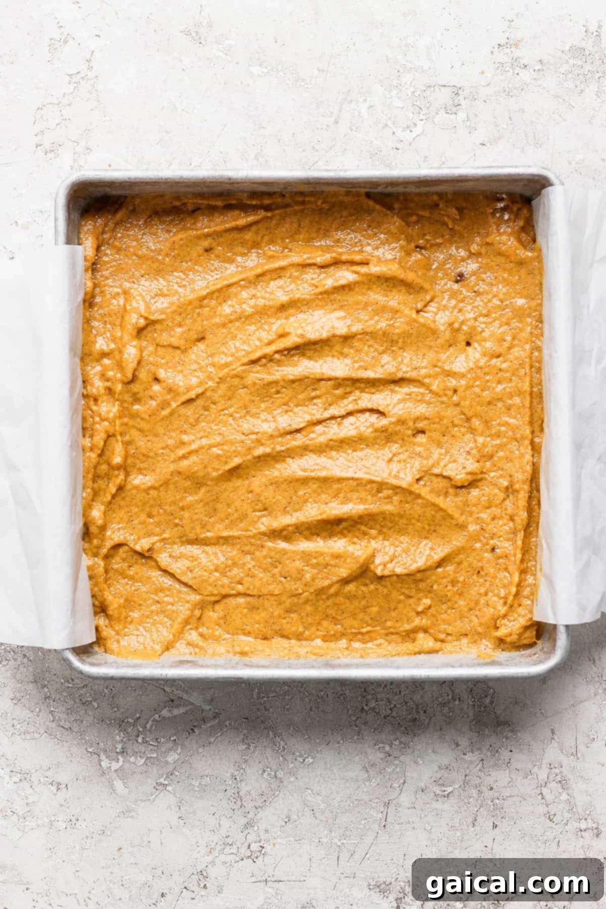 A square baking pan filled with unbaked, smooth pumpkin batter, ready for the oven. The edges of the parchment paper are visible extending from the pan.