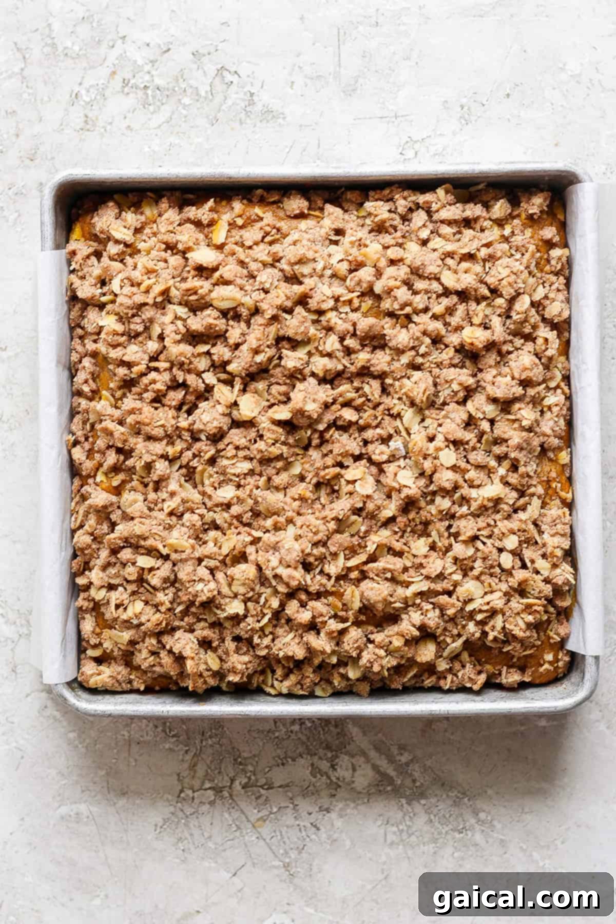 A perfectly baked square pumpkin coffee cake, glistening with a golden-brown oat and cinnamon streusel topping, still in its parchment-lined metal pan.