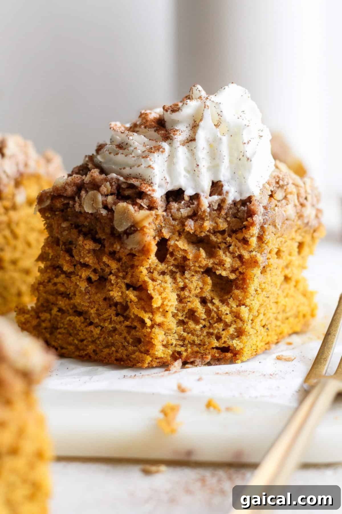 A close-up view of a scrumptious piece of pumpkin coffee cake, generously topped with streusel and a swirl of whipped cream, artfully presented on a white surface next to a gold fork.