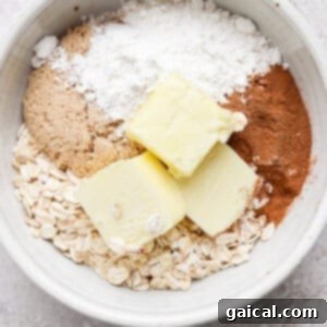 A white bowl containing oats, brown sugar, flour, cinnamon, and two slabs of butter on a light-colored surface.