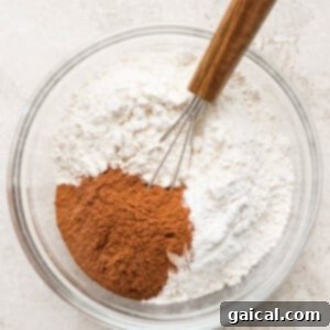 A clear glass bowl containing flour, cinnamon, and a small whisk with a wooden handle on a light-colored surface.