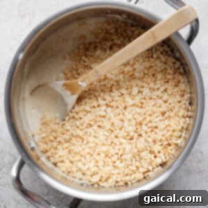 A pot containing a mixture of melted ingredients and rice cereal being stirred with a wooden spoon on a gray surface.