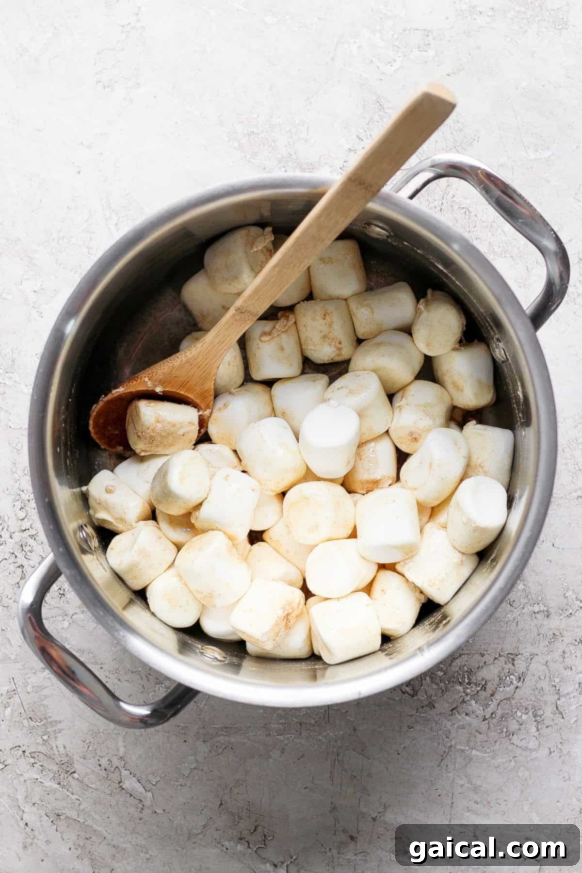 Aromatic Chai & Nutty Brown Butter Crispy Treats 3 A pot filled with large marshmallows and a wooden spoon placed on a textured surface.