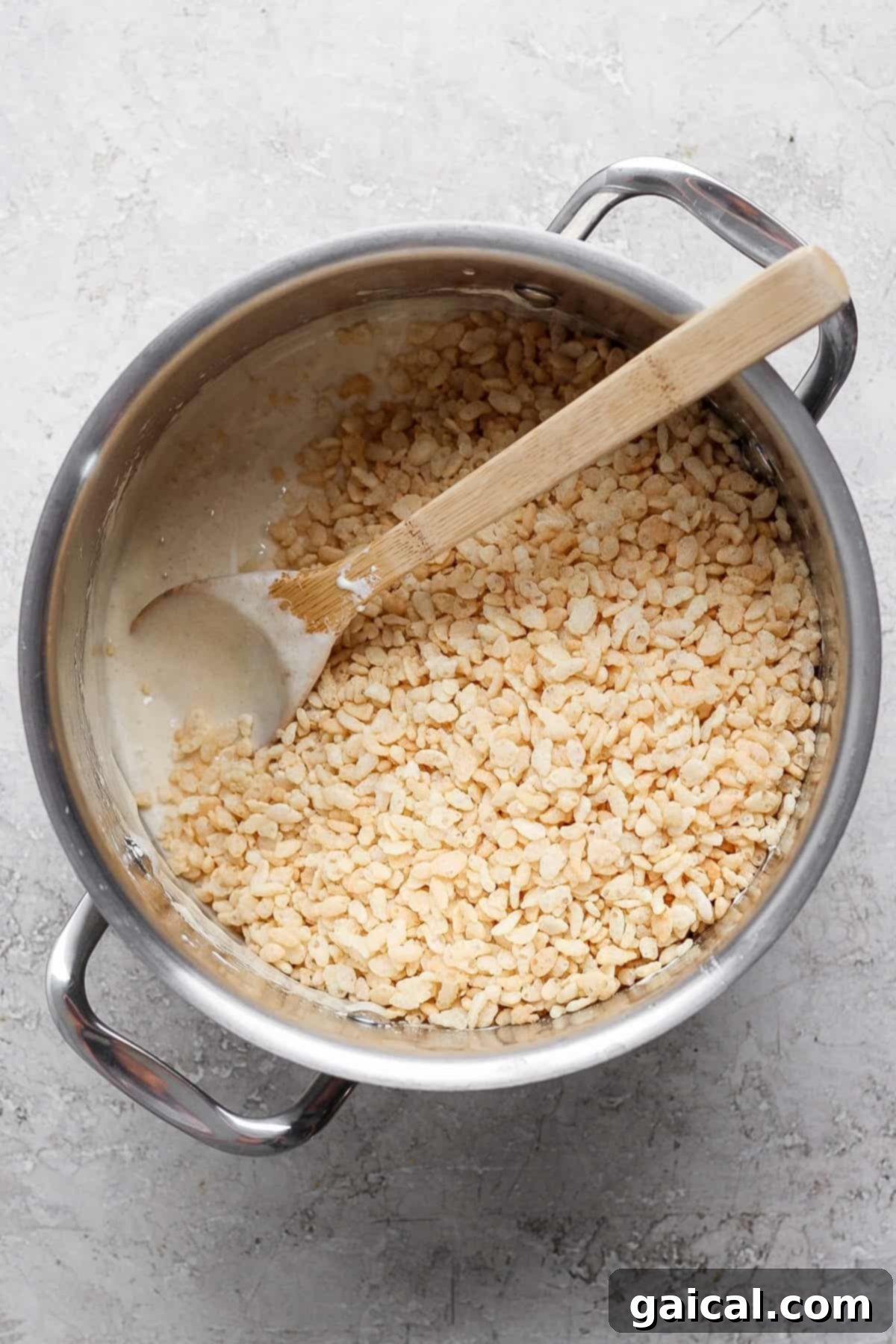 Aromatic Chai & Nutty Brown Butter Crispy Treats 4 A pot containing a mixture of melted ingredients and rice cereal being stirred with a wooden spoon on a gray surface.