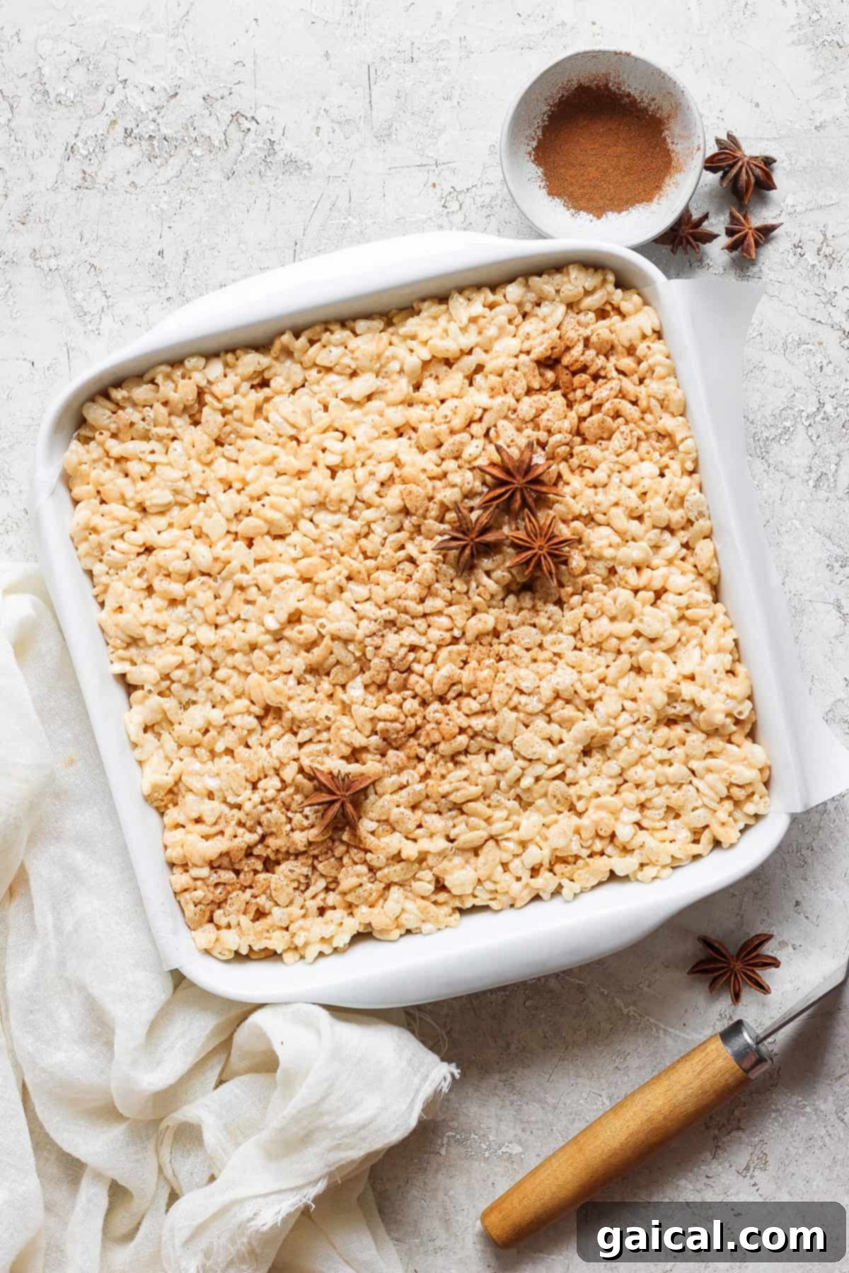 Aromatic Chai & Nutty Brown Butter Crispy Treats 6 A square white baking dish filled with rice crispy treats, garnished with star anise and cinnamon powder in the top right corner. A small bowl with cinnamon powder is beside the dish on the counter.