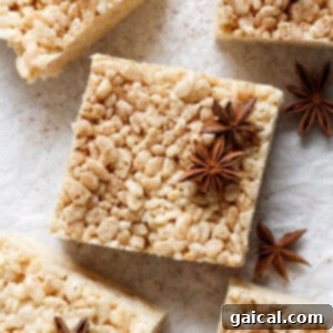 A square rice crispy treat topped with star anise spices, placed on a light-colored surface. Other identical treats and star anise pieces are shown around it.