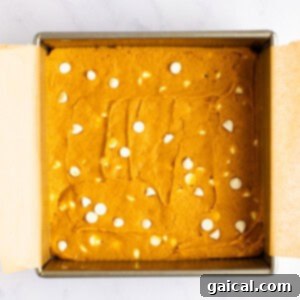 Warm Spiced Pumpkin Blondies 10 A square baking pan filled with unbaked batter topped with scattered white chocolate chips, lined with parchment paper on a marble countertop.