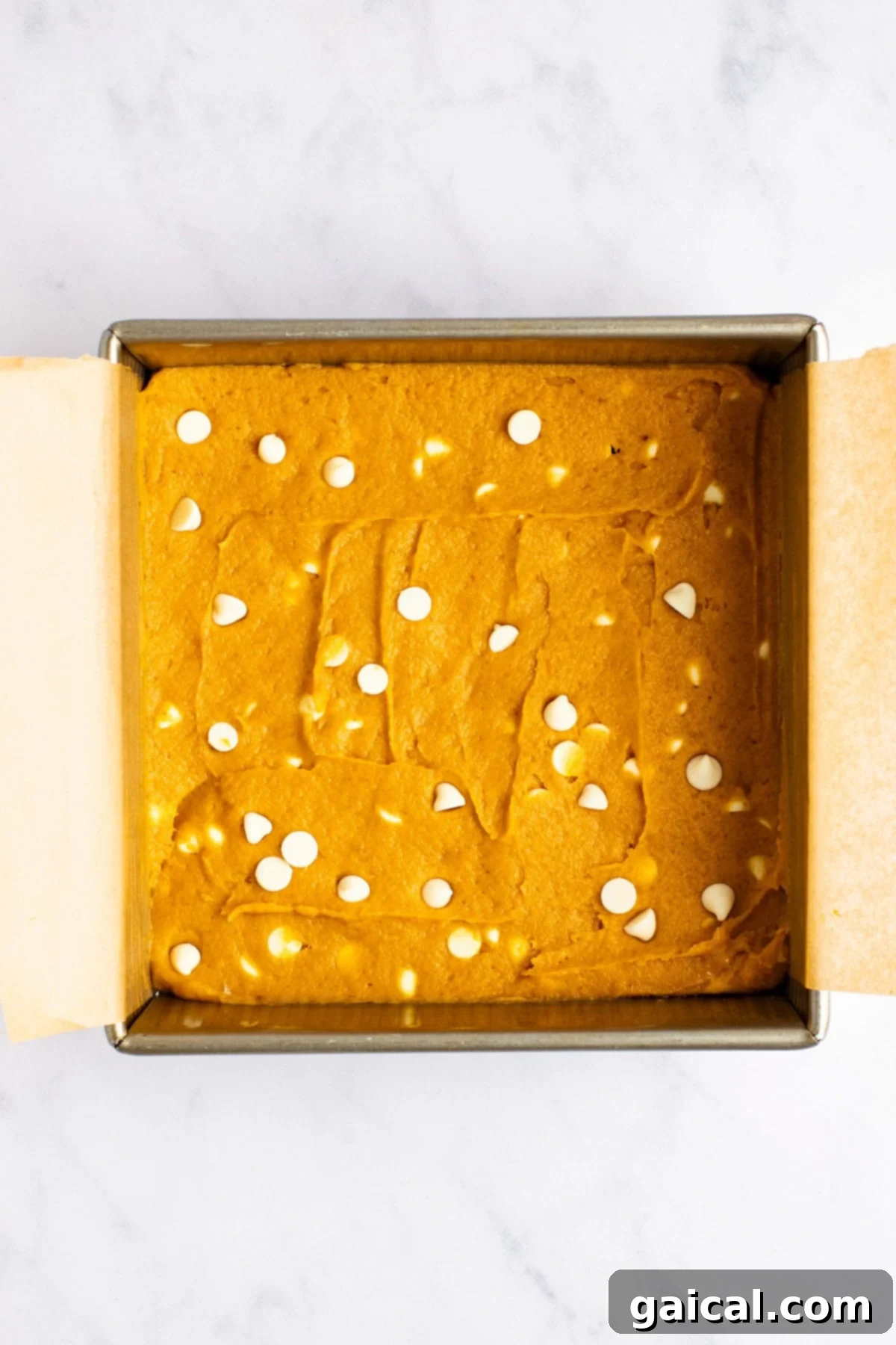 Warm Spiced Pumpkin Blondies 3 A square baking pan filled with unbaked batter topped with scattered white chocolate chips, lined with parchment paper on a marble countertop.
