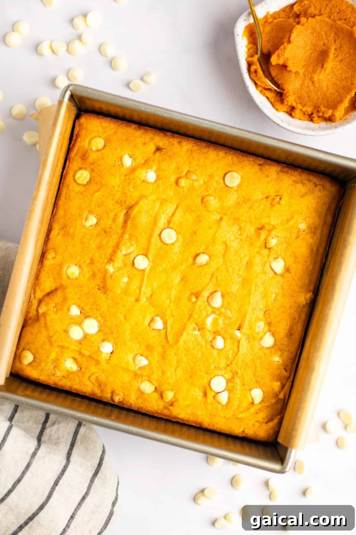 Warm Spiced Pumpkin Blondies 4 A square pan of orange-colored baked goods with white chocolate chips. A small bowl of pumpkin puree and a spoon are placed beside the pan. A striped cloth and scattered chocolate chips are also visible.