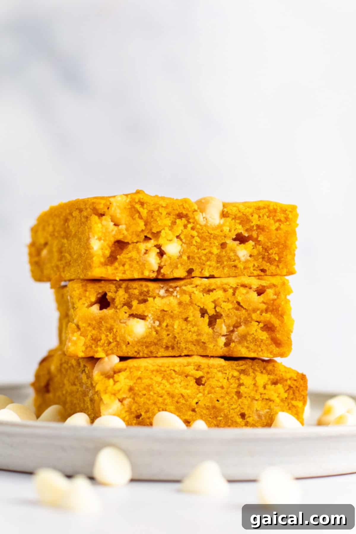 Warm Spiced Pumpkin Blondies 5 A stack of three pumpkin-flavored blondies with white chocolate chips sits on a grey plate, surrounded by scattered white chocolate chips, against a white background.