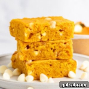 Warm Spiced Pumpkin Blondies 6 A stack of three pumpkin blondies with white chocolate chips on a white plate. Scattered white chocolate chips surround the stack.