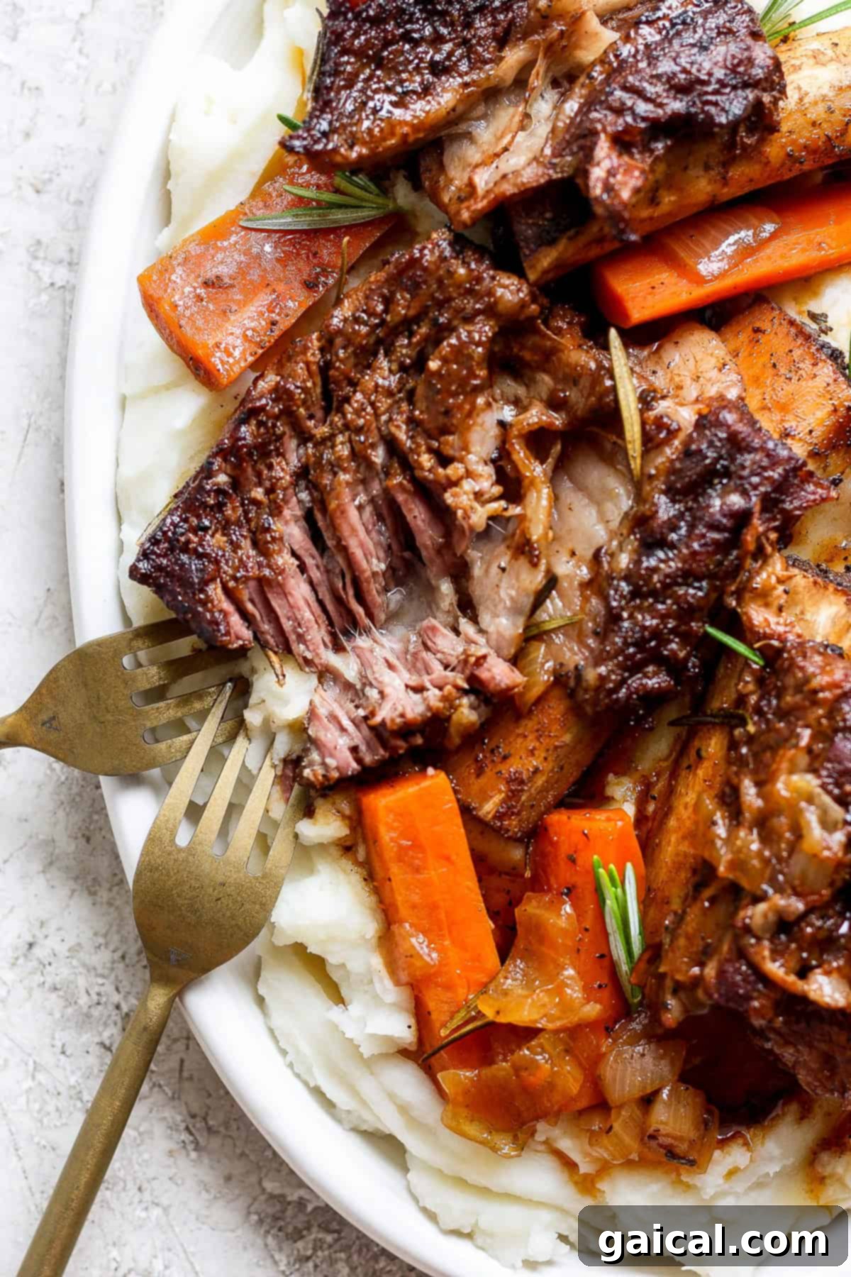 Melt-in-Your-Mouth Short Ribs 2 A plate of tender braised beef short ribs with carrots, garnished with rosemary, served on a bed of creamy mashed potatoes. Two forks are placed beside the dish.