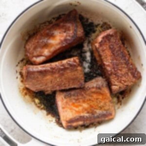 Four browned meat pieces are in a white, enameled Dutch oven on a light surface.