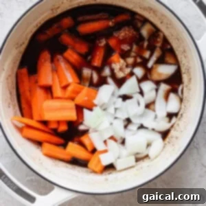 A white pot on a light surface contains chopped carrots, diced onions, minced garlic, and a dark liquid. The ingredients appear to be in the initial stages of cooking.