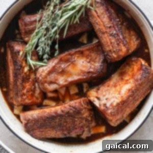 A white pot contains several pieces of cooked beef short ribs garnished with fresh rosemary and thyme.