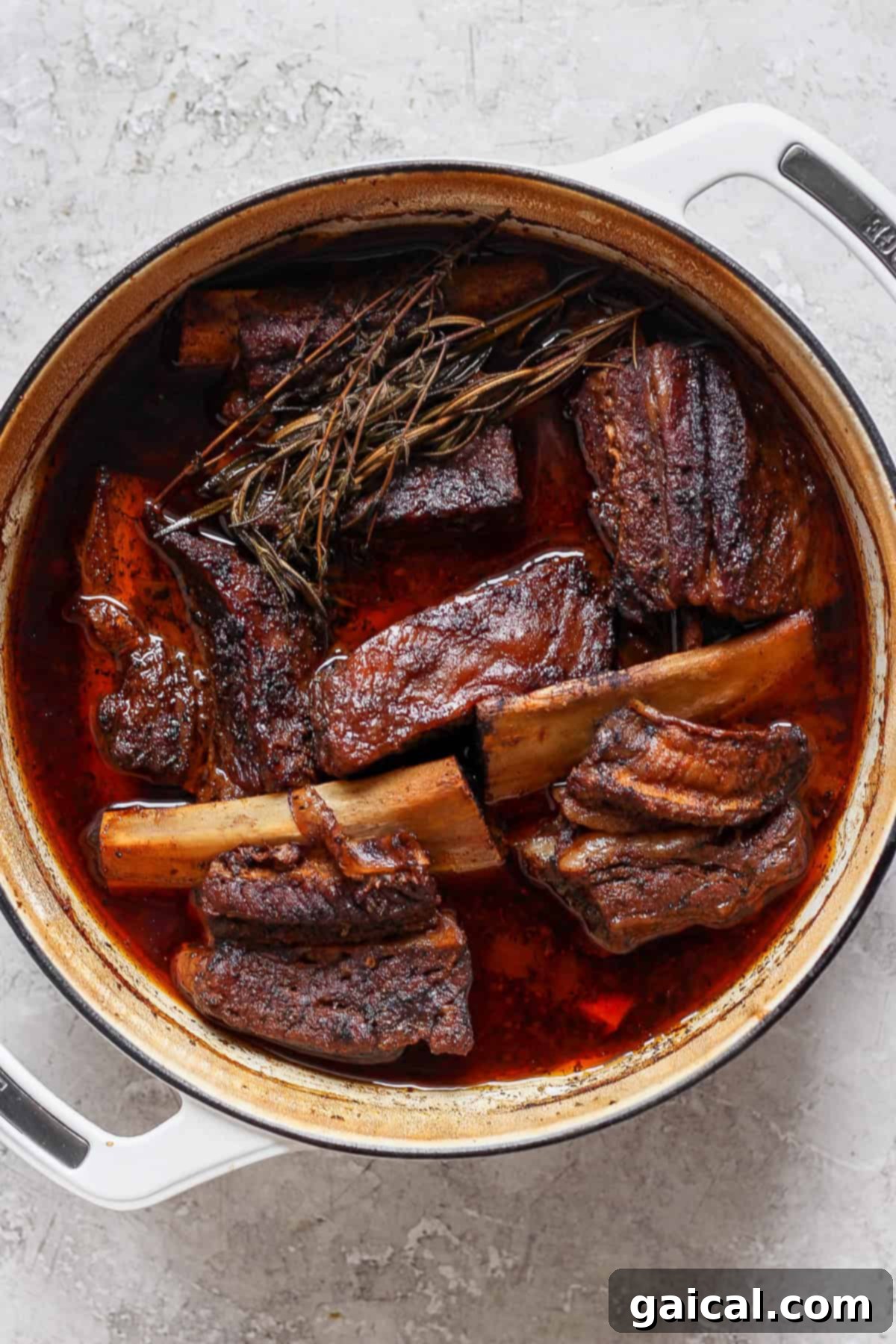 Melt-in-Your-Mouth Short Ribs 4 A large pot filled with braised short ribs in a rich, dark sauce, garnished with herbs on a light gray surface.