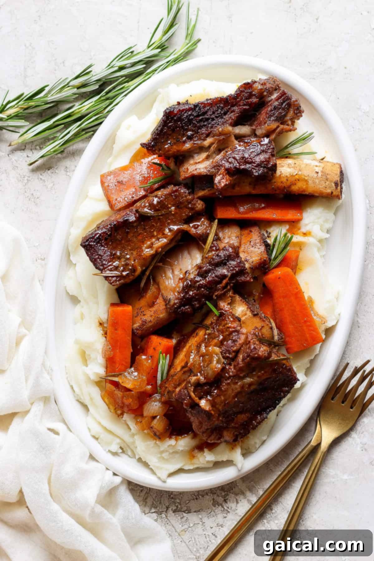 Melt-in-Your-Mouth Short Ribs 6 A white oval plate with mashed potatoes, roasted carrot slices, and beef short ribs garnished with fresh rosemary sprigs. A fork and knife are placed beside the plate on a white surface.