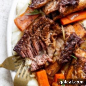 A plate of tender roasted meat with a crispy exterior, served on a bed of mashed potatoes and garnished with cooked carrots and rosemary, with two forks visible on the side.
