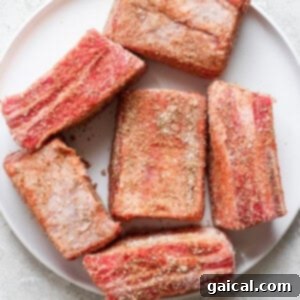 A white plate with six seasoned raw beef short ribs on a light gray surface.