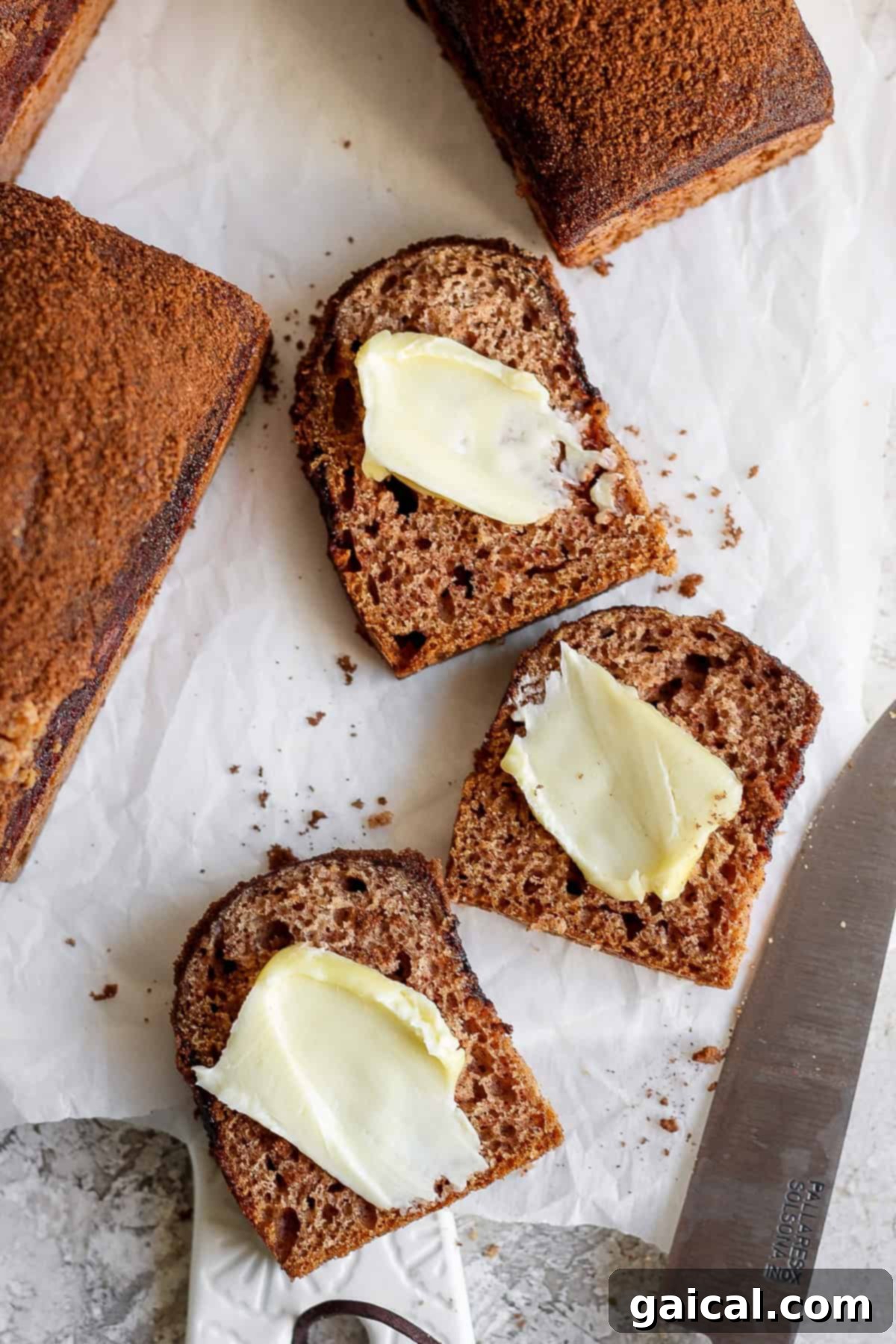 Two slices of dark bread with butter on a parchment paper, next to a knife with a partially visible bread loaf.