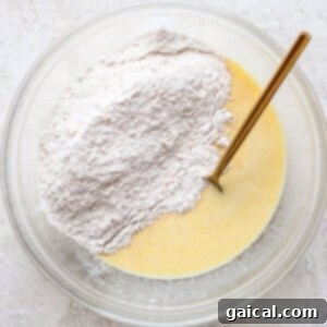 A clear bowl contains a mixture of liquid ingredients and dry flour with a gold spoon, set on a textured white surface.
