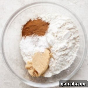 A glass bowl containing flour, brown sugar, cinnamon, baking powder, and baking soda, on a light textured surface.