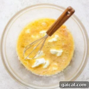 A glass bowl containing a beaten egg mixture and dollops of cream or butter, with a whisk resting inside.