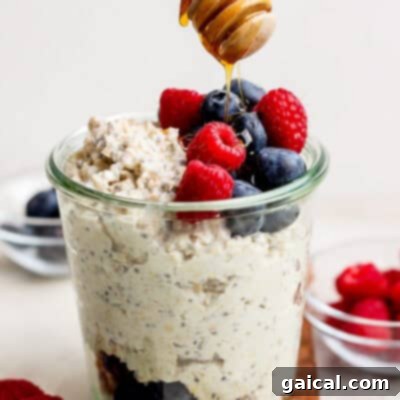 A glass jar with layers of oatmeal, fruit, and a creamy mixture, topped with berries and chia seeds, next to a spoon.