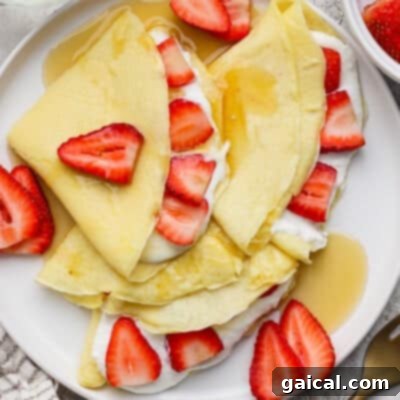 Crepes folded and topped with sliced strawberries, cream, and syrup on a white plate.