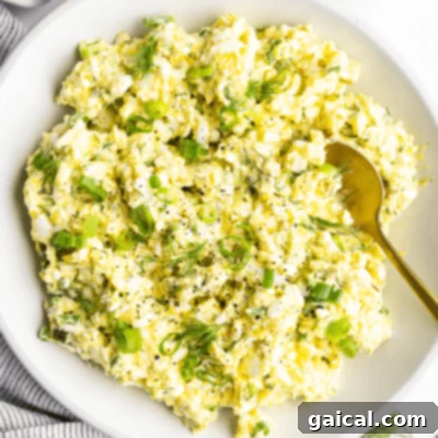 A bowl of egg salad with chopped green onions and dill, accompanied by a gold spoon.