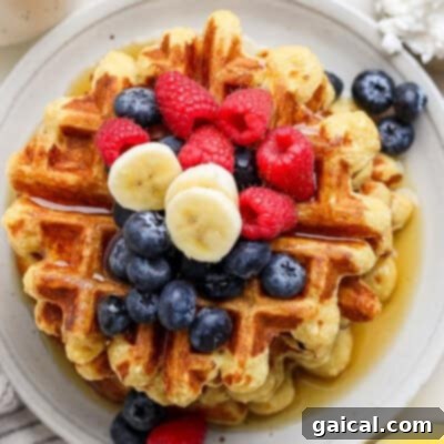 A stack of waffles topped with blueberries, raspberries, banana slices, and drizzled with syrup on a round white plate.